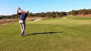 Professional golfer mid-swing on manicured fairway with pristine green grass, clear blue sky, and lush landscaping in background, no people watching, serene golf course environment